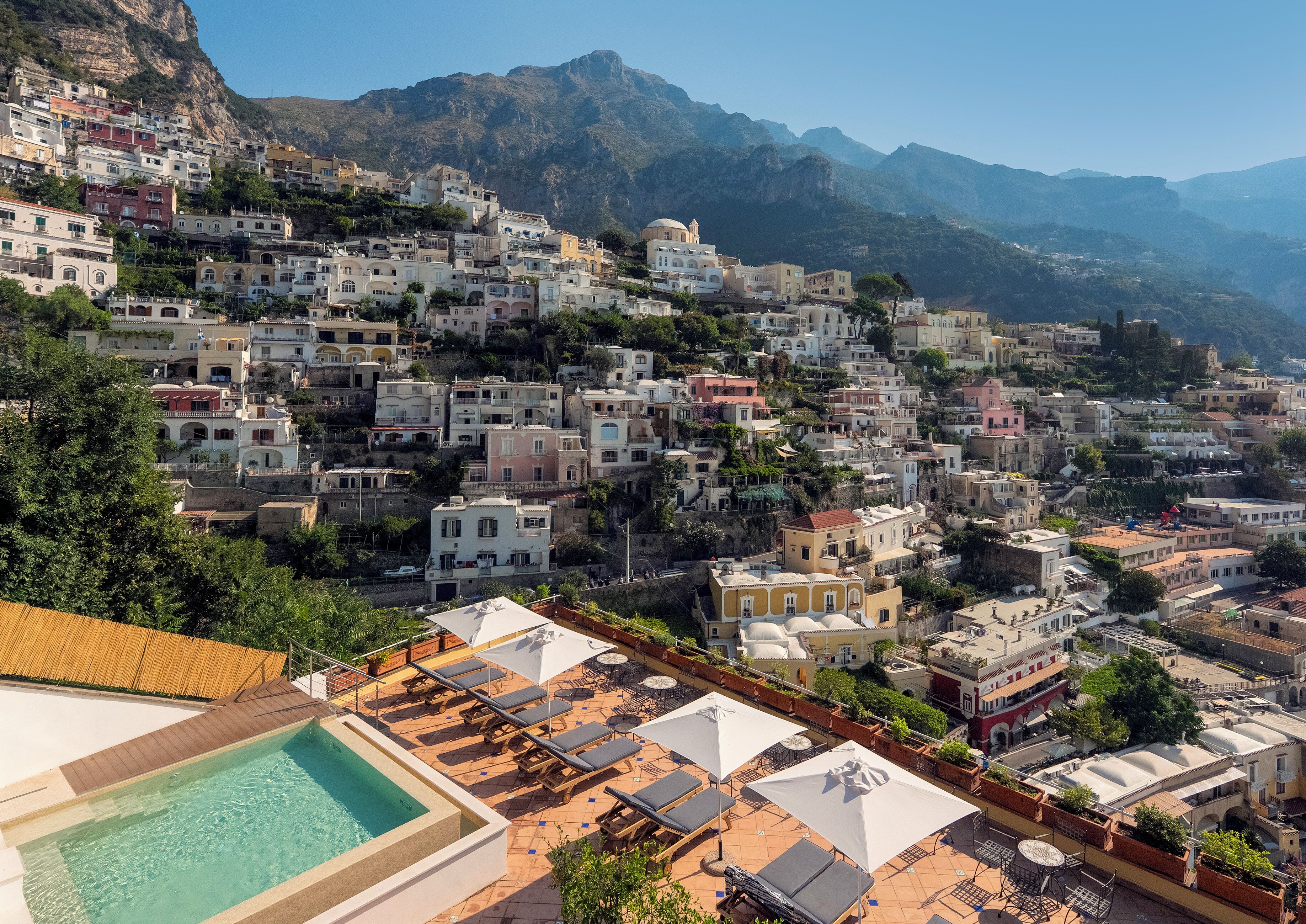 A hillside town with terraced buildings, a pool area with sun loungers, and a mountain backdrop.
