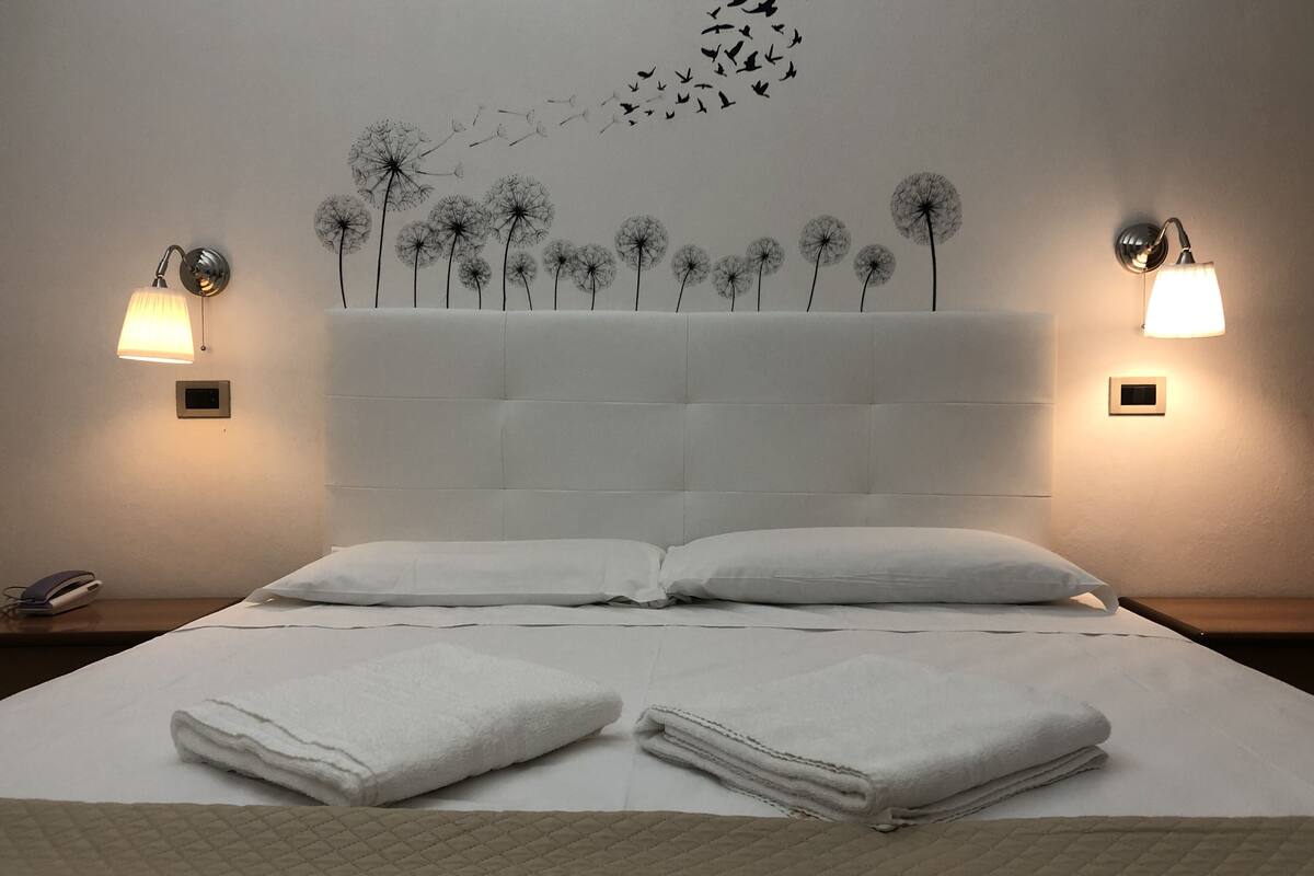 Un lit bien fait, avec deux oreillers et des serviettes pliées, une tête de lit décorée de motifs de pissenlit et d’oiseaux, et deux lampes fixées au mur, de part et d’autre du lit.
