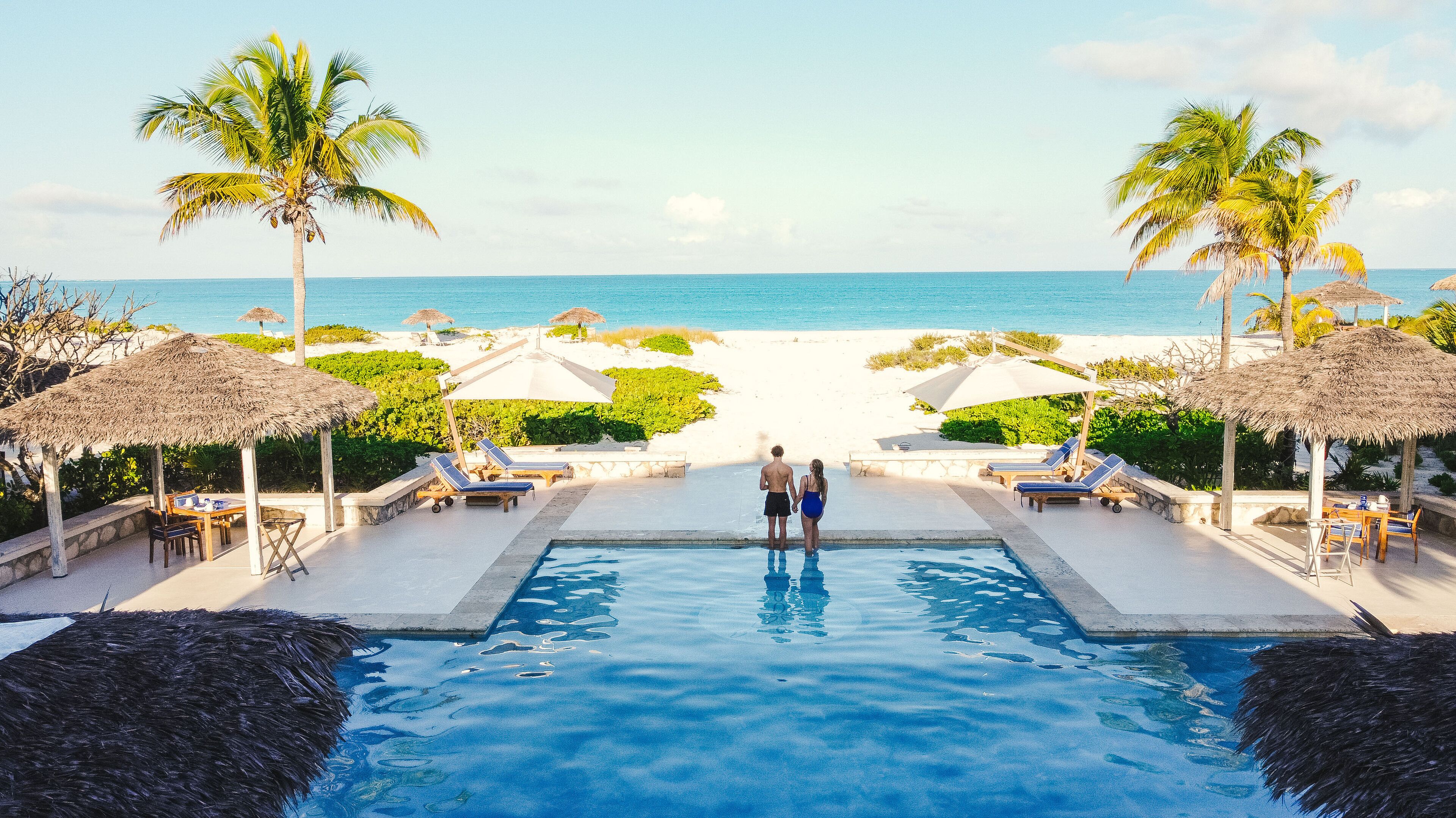A pool area with lounge chairs and umbrellas, leading to a beach with palm trees.