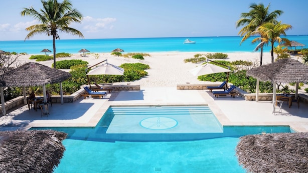 A pool area with a view of the beach and ocean, surrounded by palm trees and thatched umbrellas. - Pine Cay, Turks and Caicos (Pine Cay)