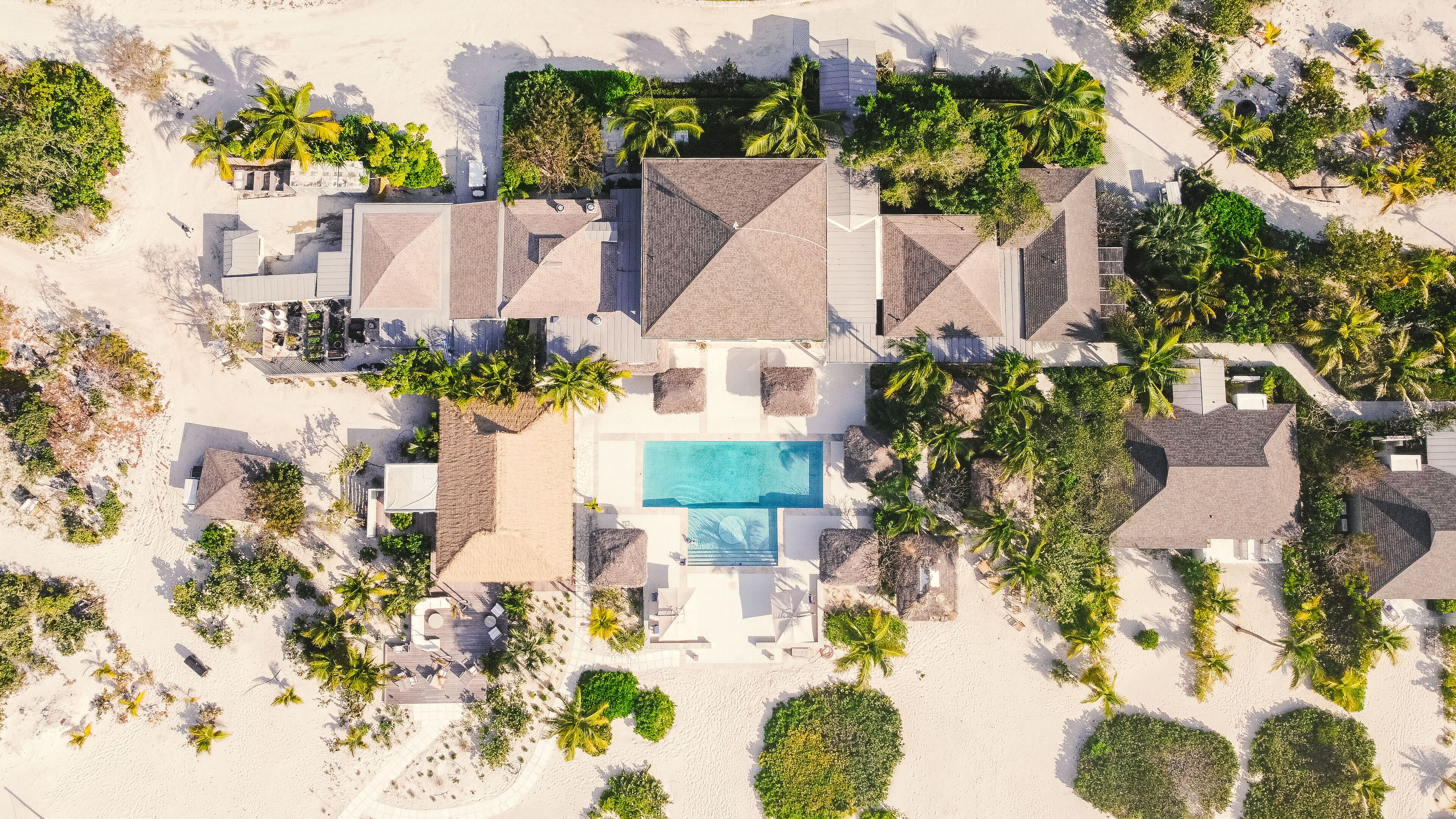 An aerial view of a resort with a central swimming pool, multiple villas, and surrounding palm trees.