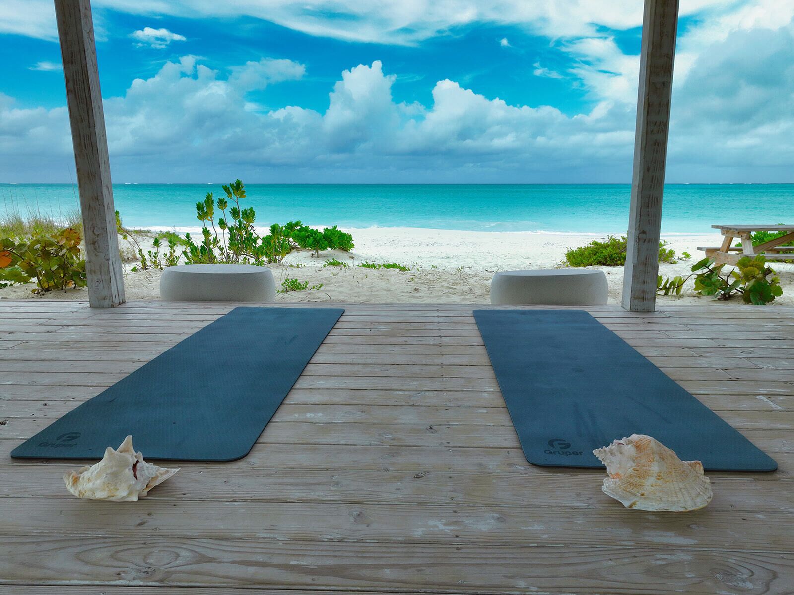 A wooden deck with two black yoga mats and seashells, overlooking a beach with white sand and turquoise water.