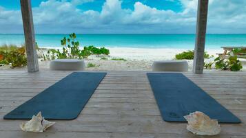 A wooden deck with two black yoga mats and seashells, overlooking a beach with white sand and turquoise water.
