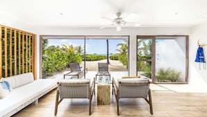 A modern living room with a sofa, two wicker chairs, a wooden coffee table, and large glass doors leading to a tropical garden and ocean view. - Pine Cay, Turks and Caicos (Pine Cay)