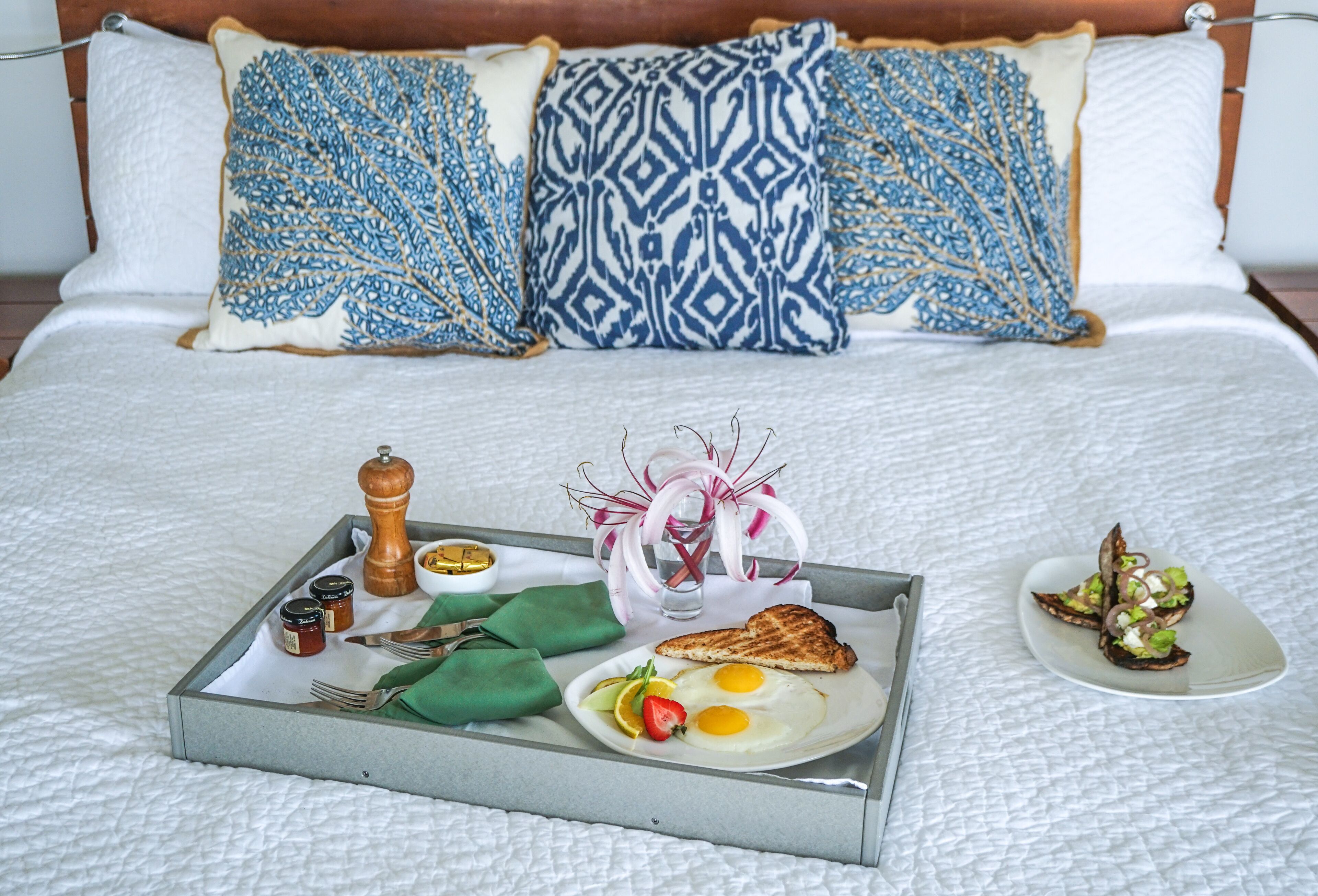 A neatly made breakfast tray with toast, eggs, fruit, and condiments on a bed with patterned pillows.