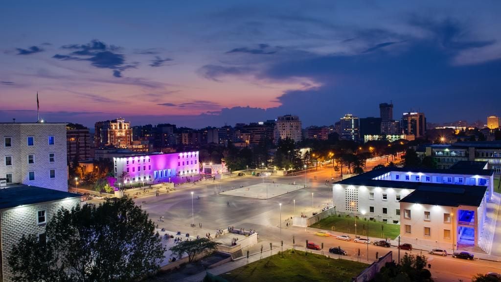 Paisatge urbà al capvespre, amb edificis il·luminats i una gran plaça oberta.