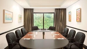 A conference room with a long table, chairs, and a view of greenery through large windows.