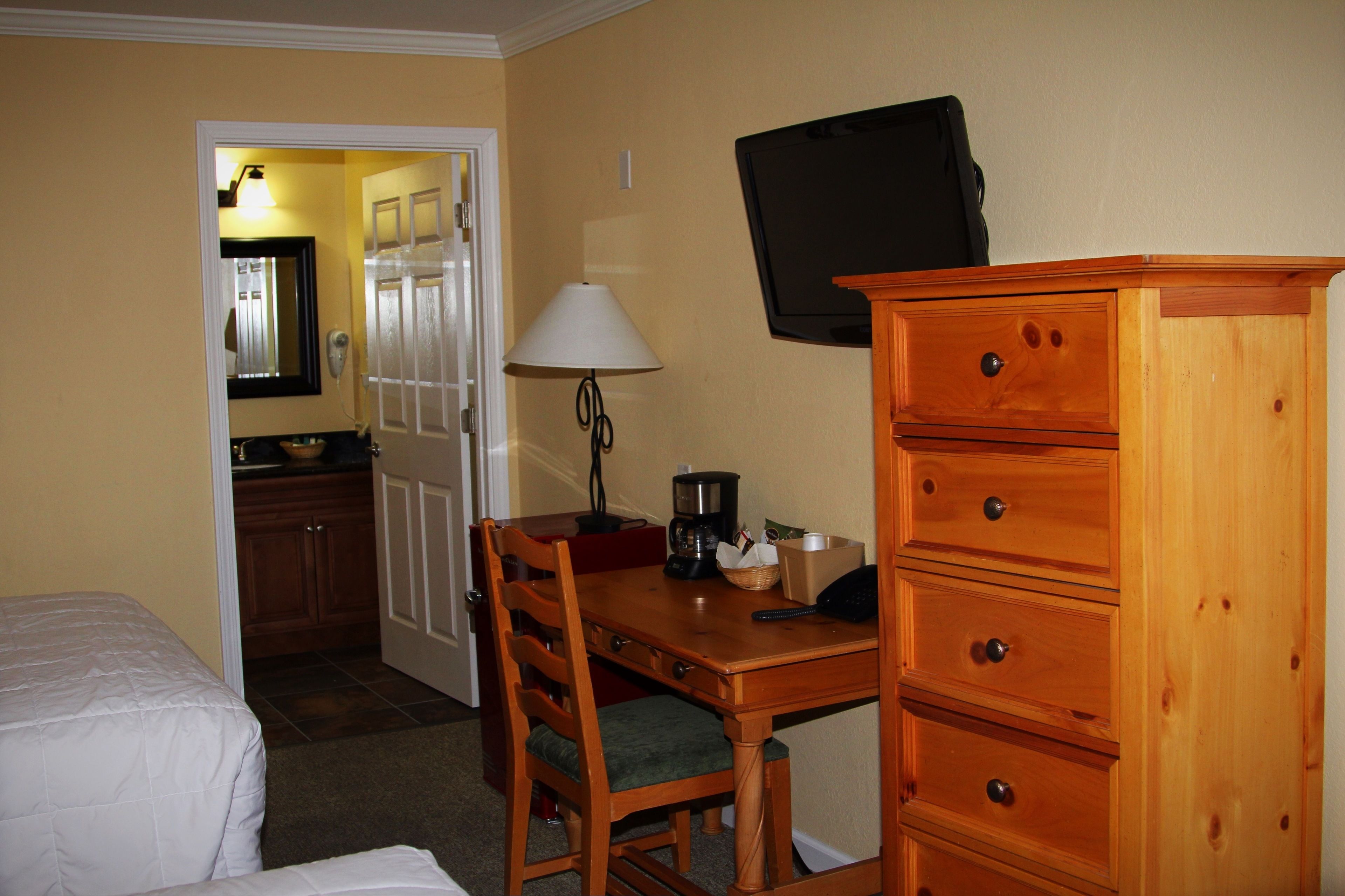 Quarto de hotel com cama, escrivaninha de madeira com luminária, televisão e cómoda.