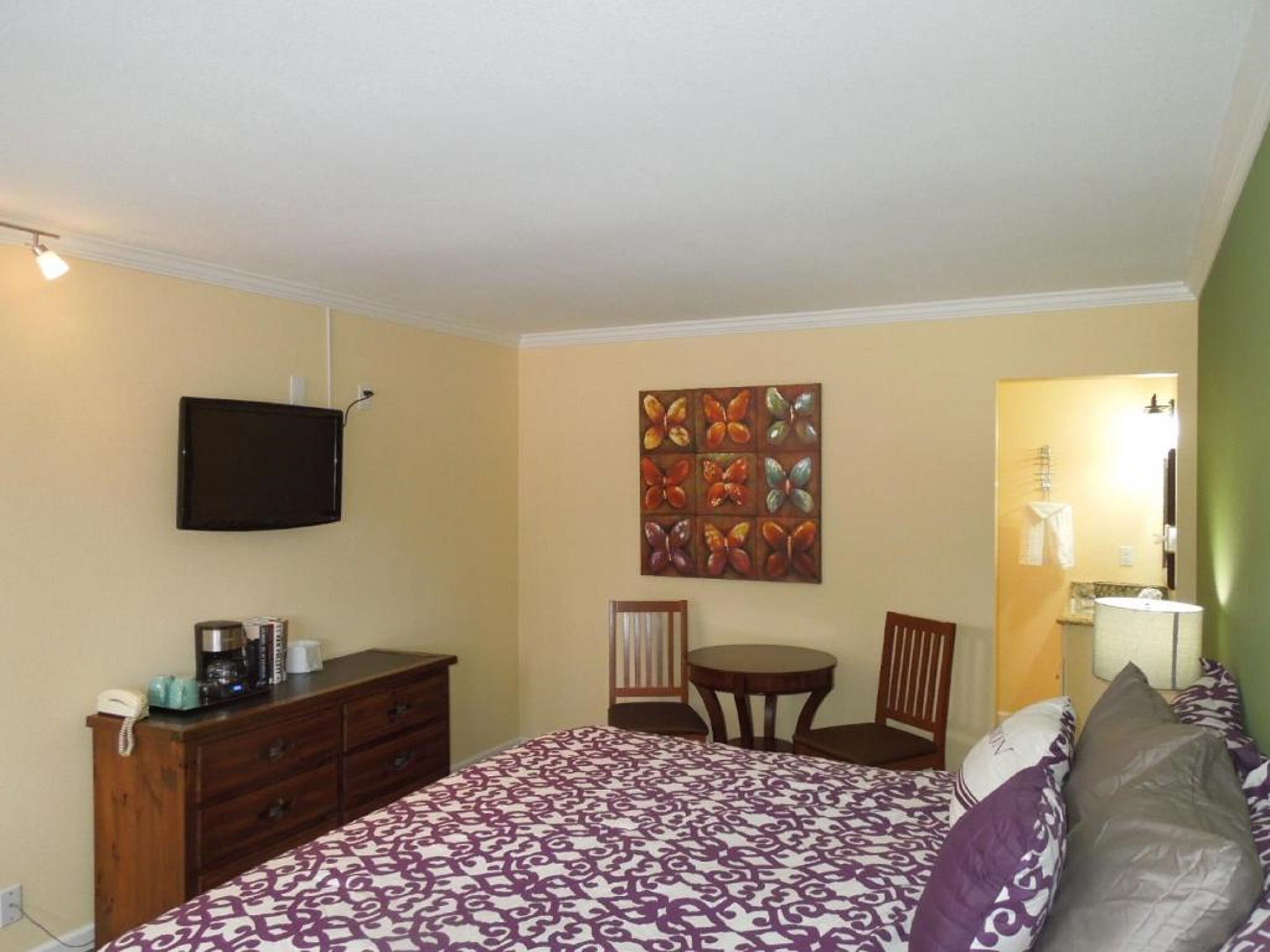 Una camera d'albergo con un letto, una cassettiera, una TV, un quadro, una sedia, un tavolo e una lampada.