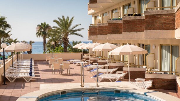 Zona de piscina con sillones reclinables y sombrillas, vista despejada al mar, y un edificio con balcones.