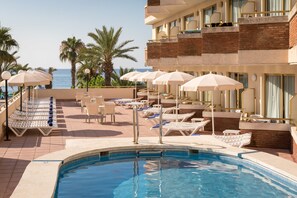 A pool area with lounge chairs and umbrellas, a clear view of the sea, and a building with balconies. - htop Royal Sun (Santa Susanna)