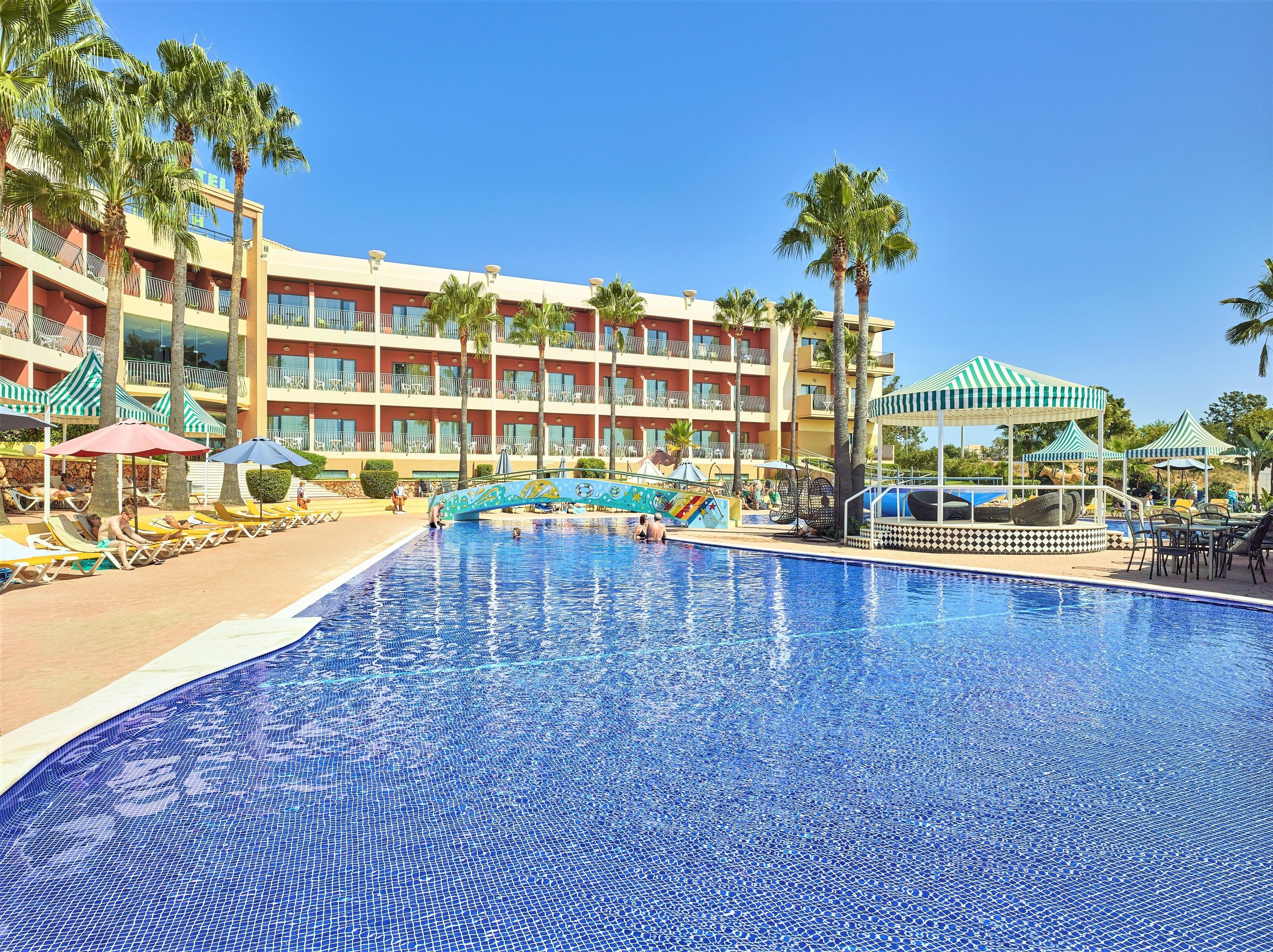 Ein Hotel mit großem Schwimmbad, Palmen und einem farbenfrohen Wandgemälde.