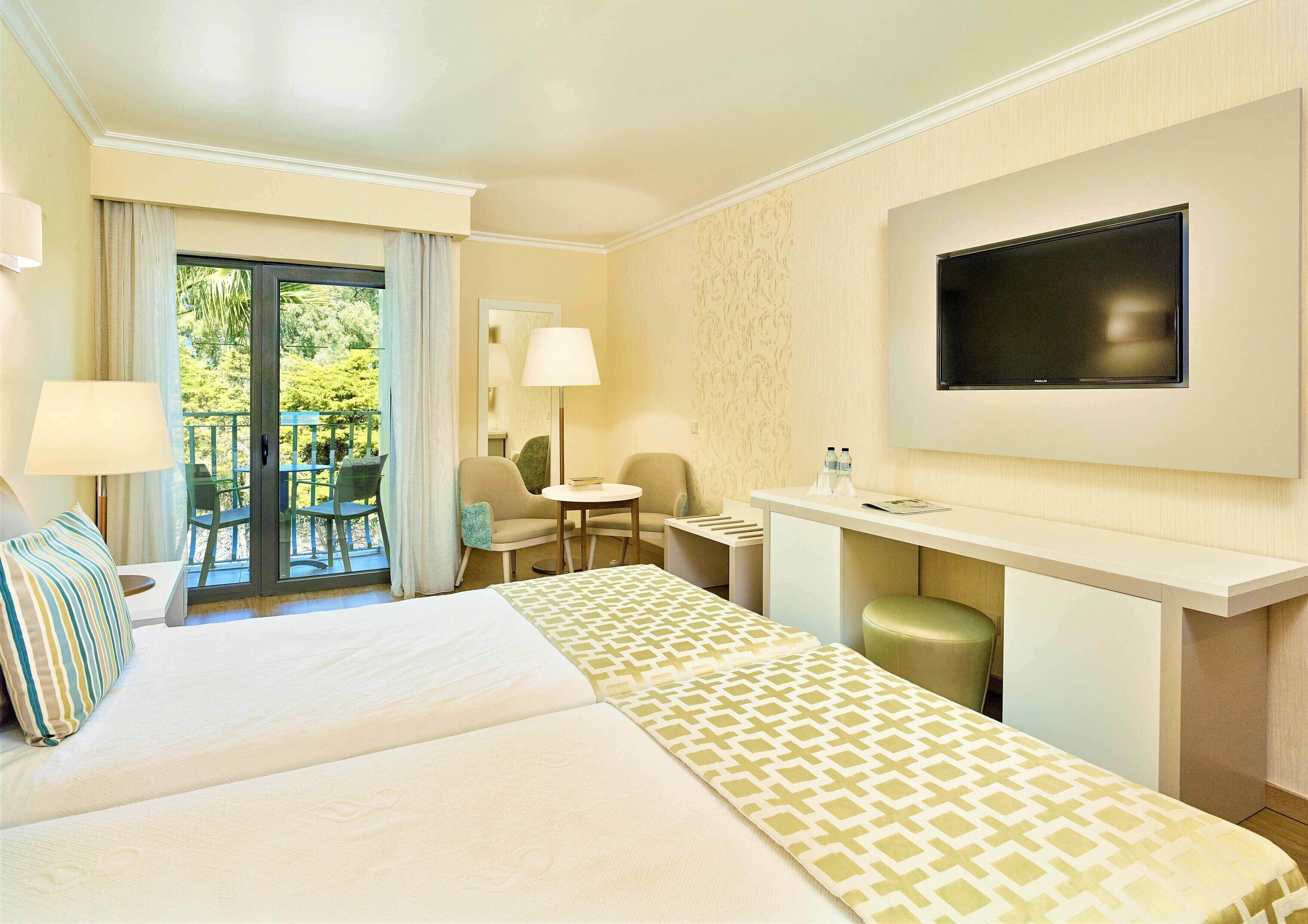 A hotel room with a bed, a desk, a chair, a TV, and a balcony with outdoor seating.