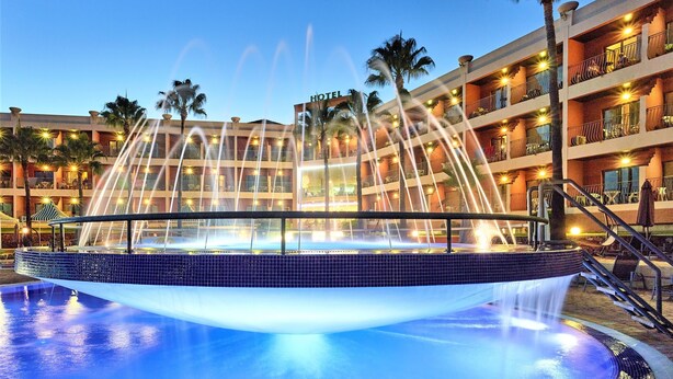 A hotel with a pool featuring a water fountain, palm trees, and a multi-story building with balconies. - Hotel Baia Grande (Albufeira)