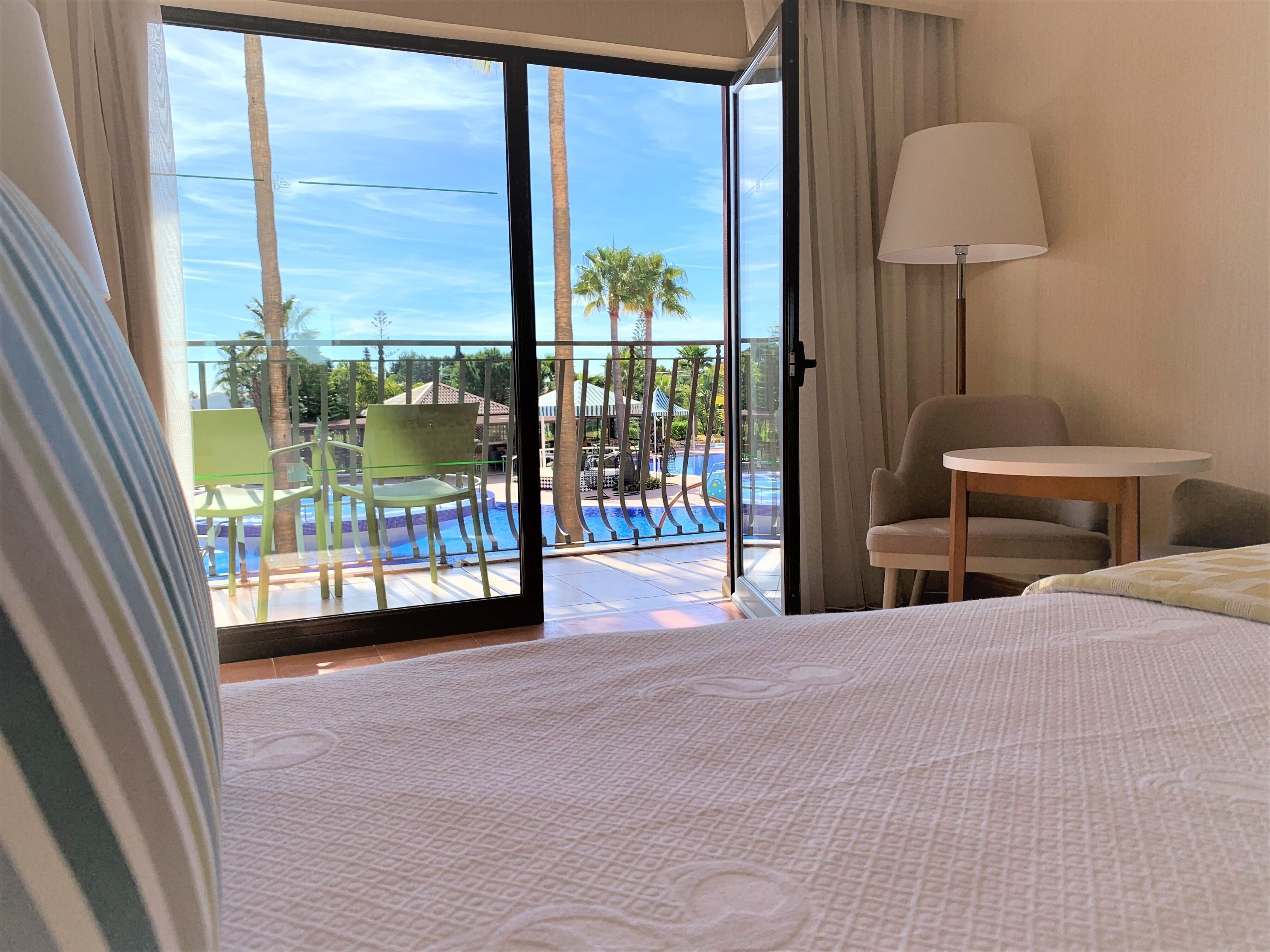 A hotel room with a bed, a chair, a table, a lamp, and a view of a pool and palm trees.