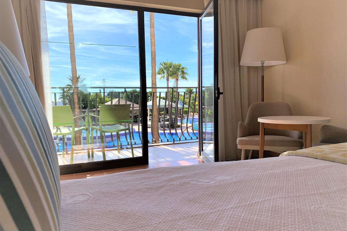 A hotel room with a bed, a chair, a table, a lamp, and a view of a pool and palm trees.