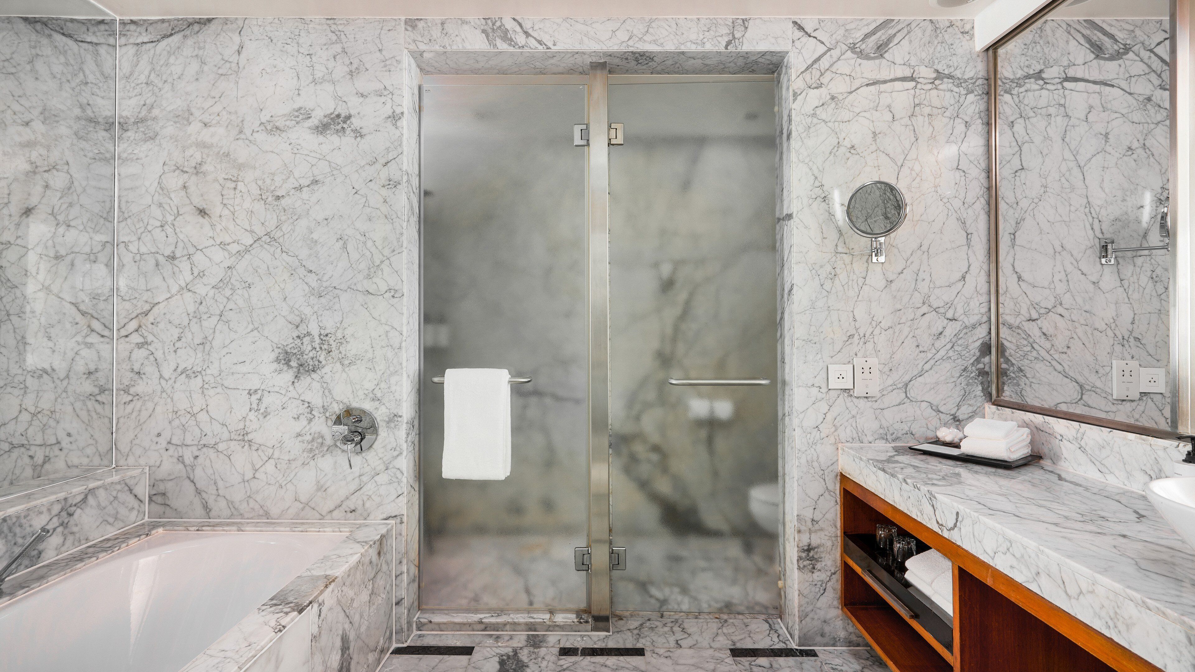 Un baño moderno con ducha de vidrio, paredes de mármol, tocador de madera y un espejo grande.