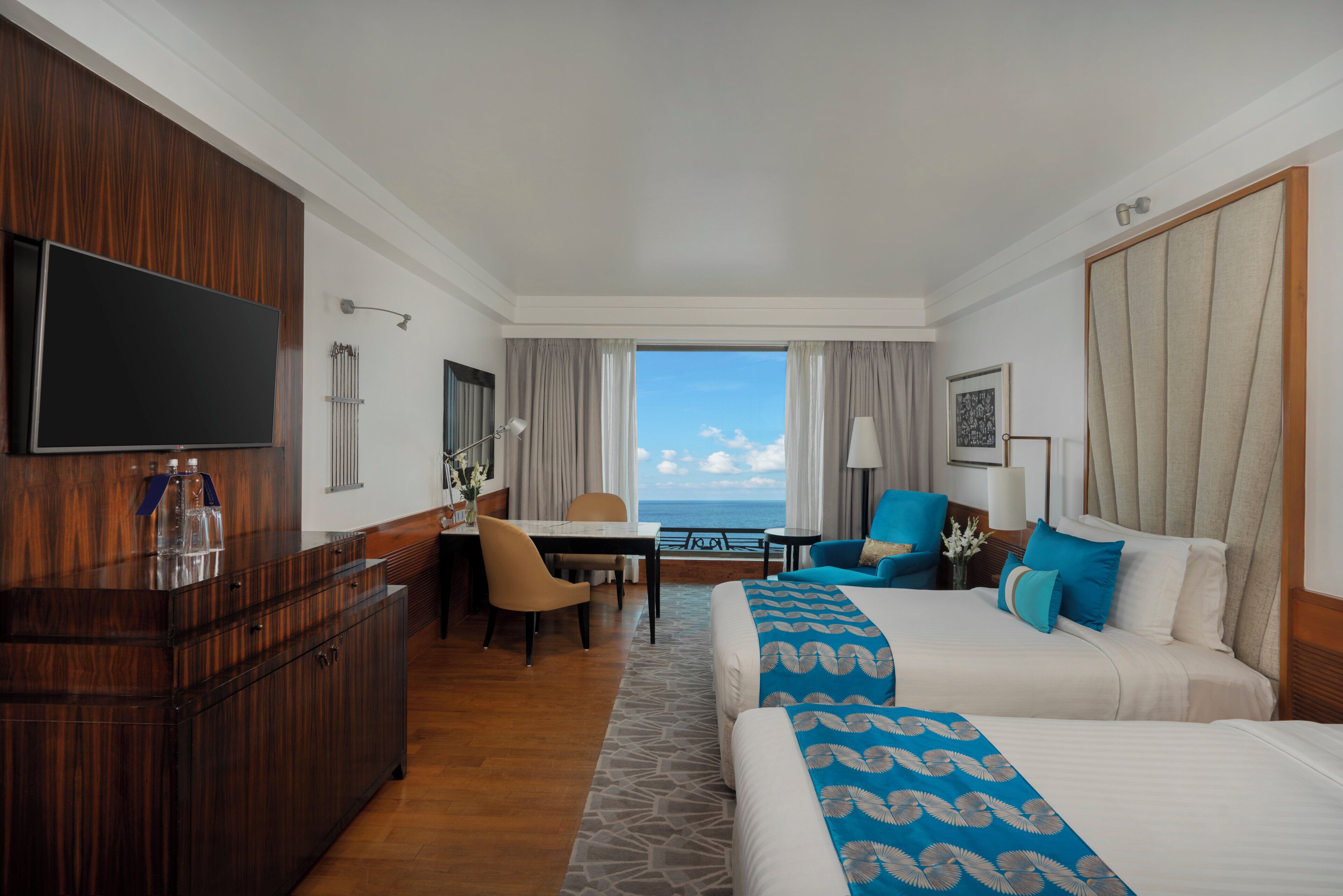 A hotel room with a large bed, a smaller bed, a desk, a chair, a television, and a view of the sea.