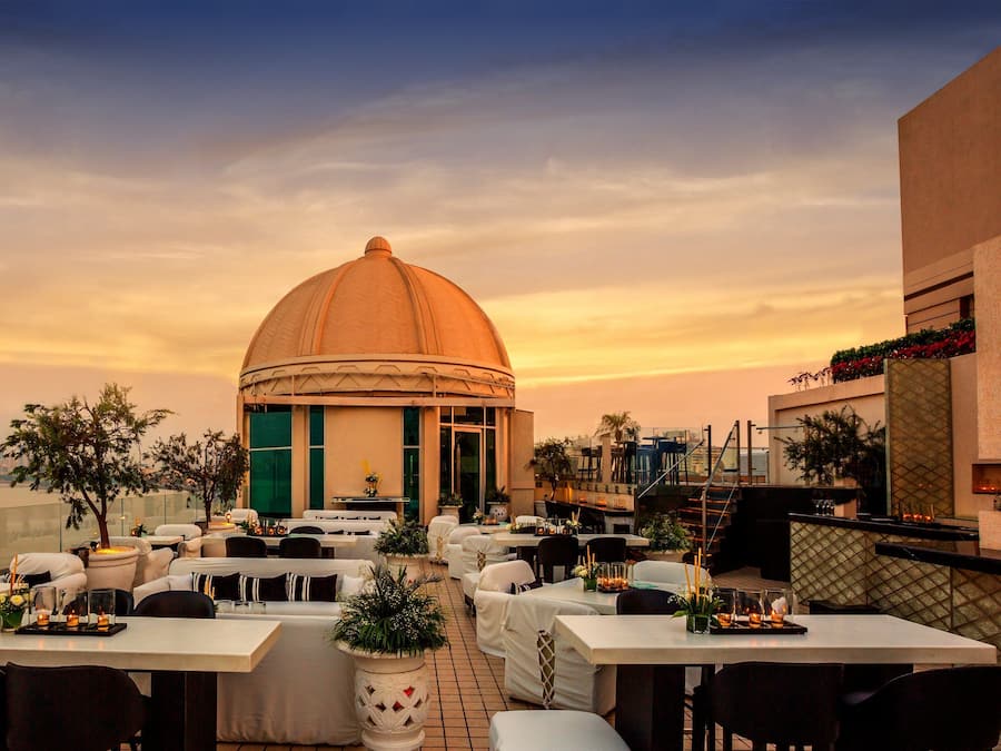 An outdoor dining area with tables and chairs, a large dome structure, and a view of the city skyline at sunset.