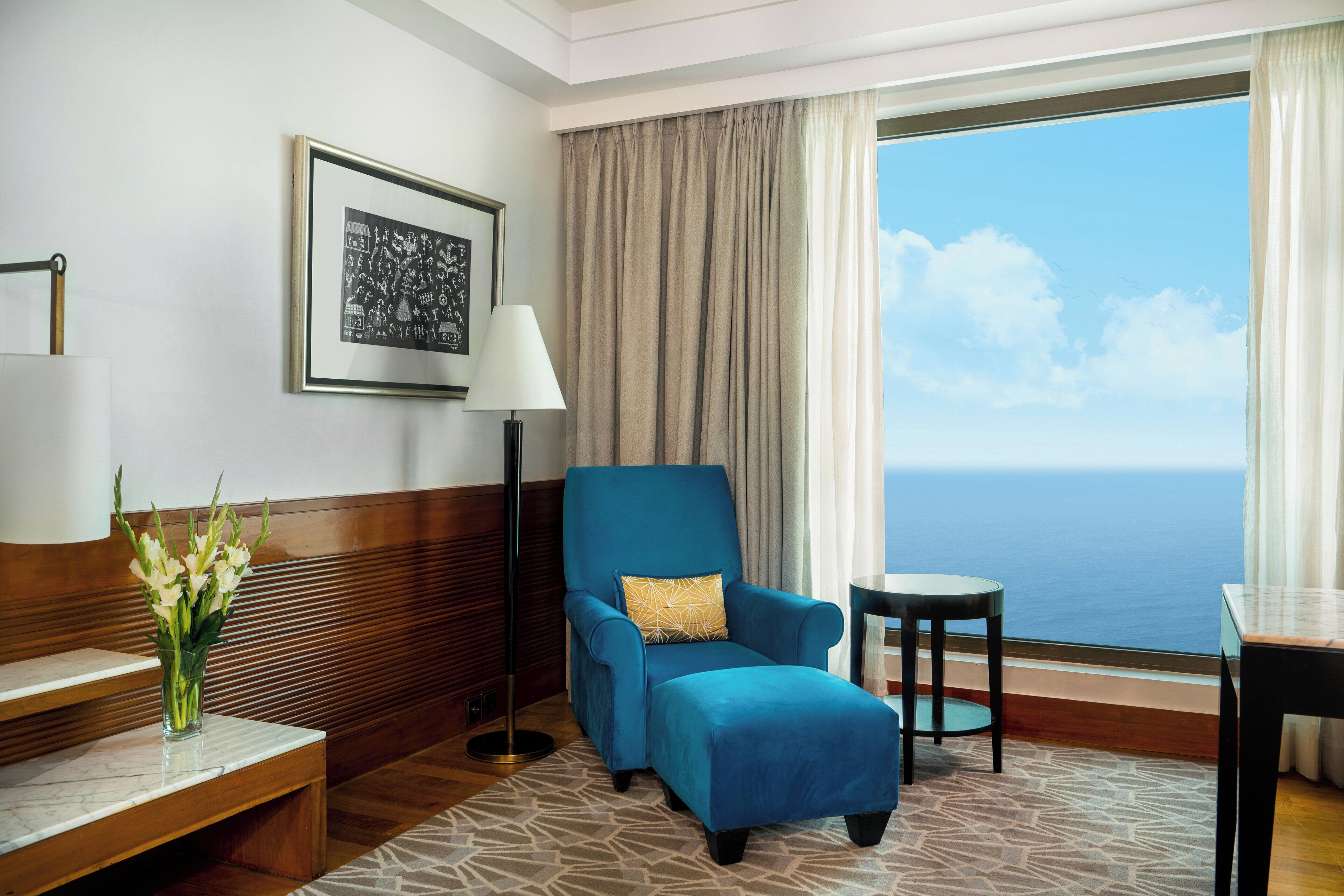 Habitación de hotel con un ventanal grande, un sillón azul con un cojín amarillo, una mesita redonda y una lámpara de pie.