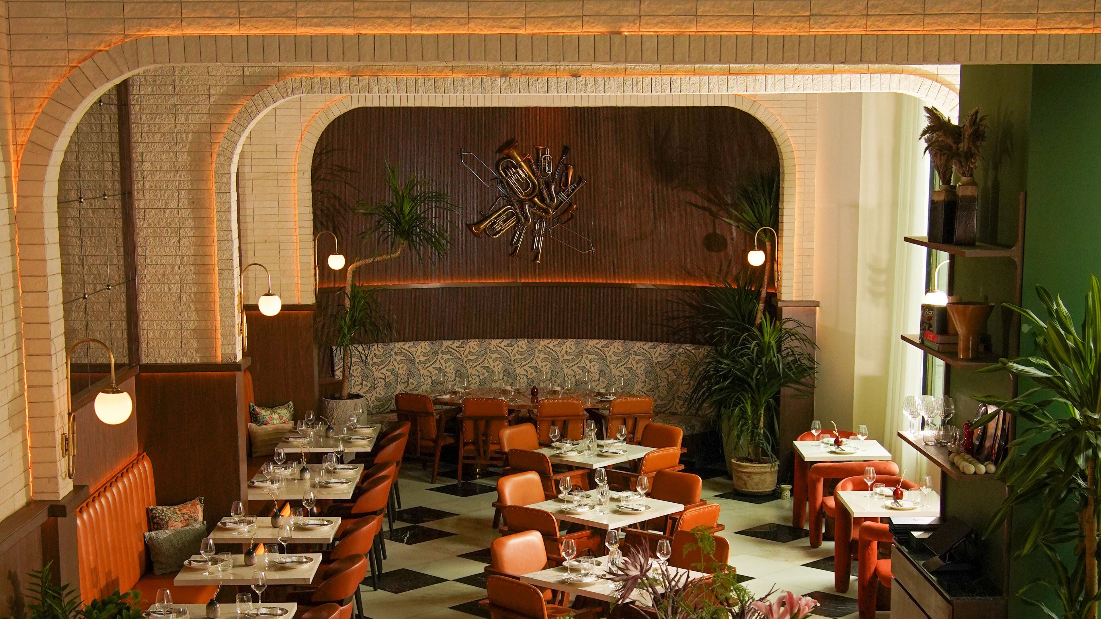 A restaurant with a checkered floor, brick arches, and orange chairs. Tables are set with wine glasses and plates.