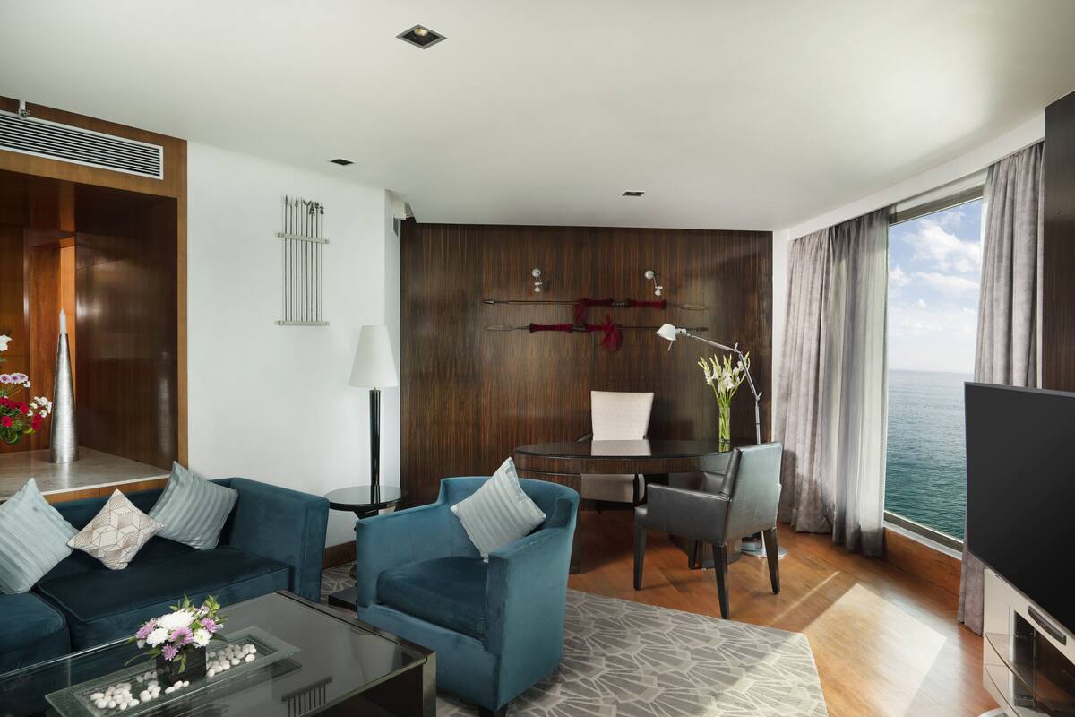 A modern hotel room with a blue sofa, a glass coffee table with flowers, a desk with a chair, a TV, and a view of the sea.