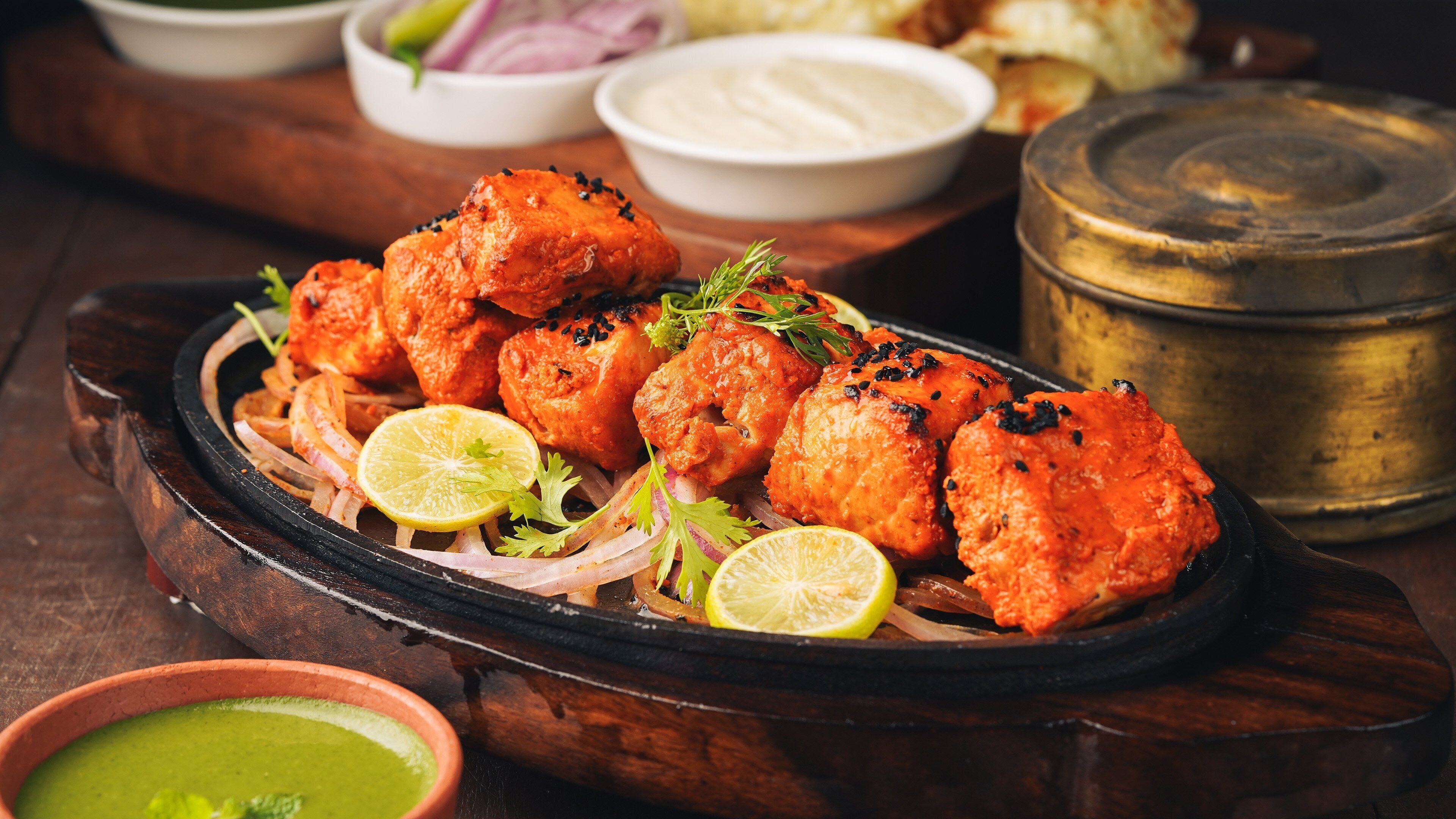 A plate of tandoori chicken with lemon slices and onions, accompanied by various sauces and a brass container.