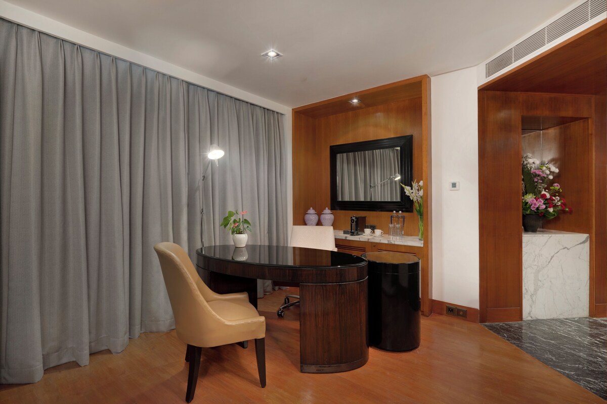 A modern hotel room with a wooden dining table, a beige chair, a small potted plant, a coffee machine, and a marble countertop.