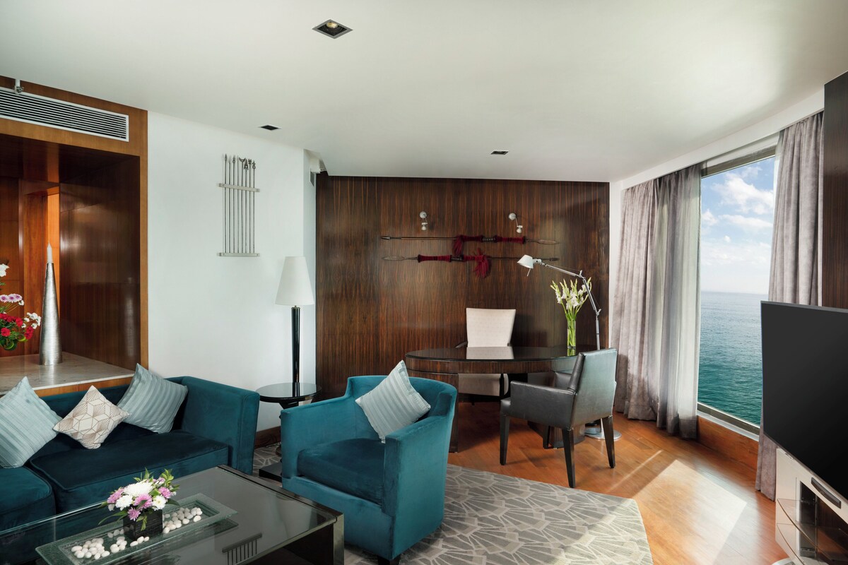 A modern hotel room with a blue sofa, a glass coffee table with flowers, a desk with a chair, a TV, and a view of the sea.