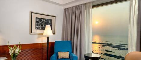 Habitación de hotel con un ventanal que ofrece vistas al mar, un sillón azul, una lámpara de pie y un cuadro en la pared.