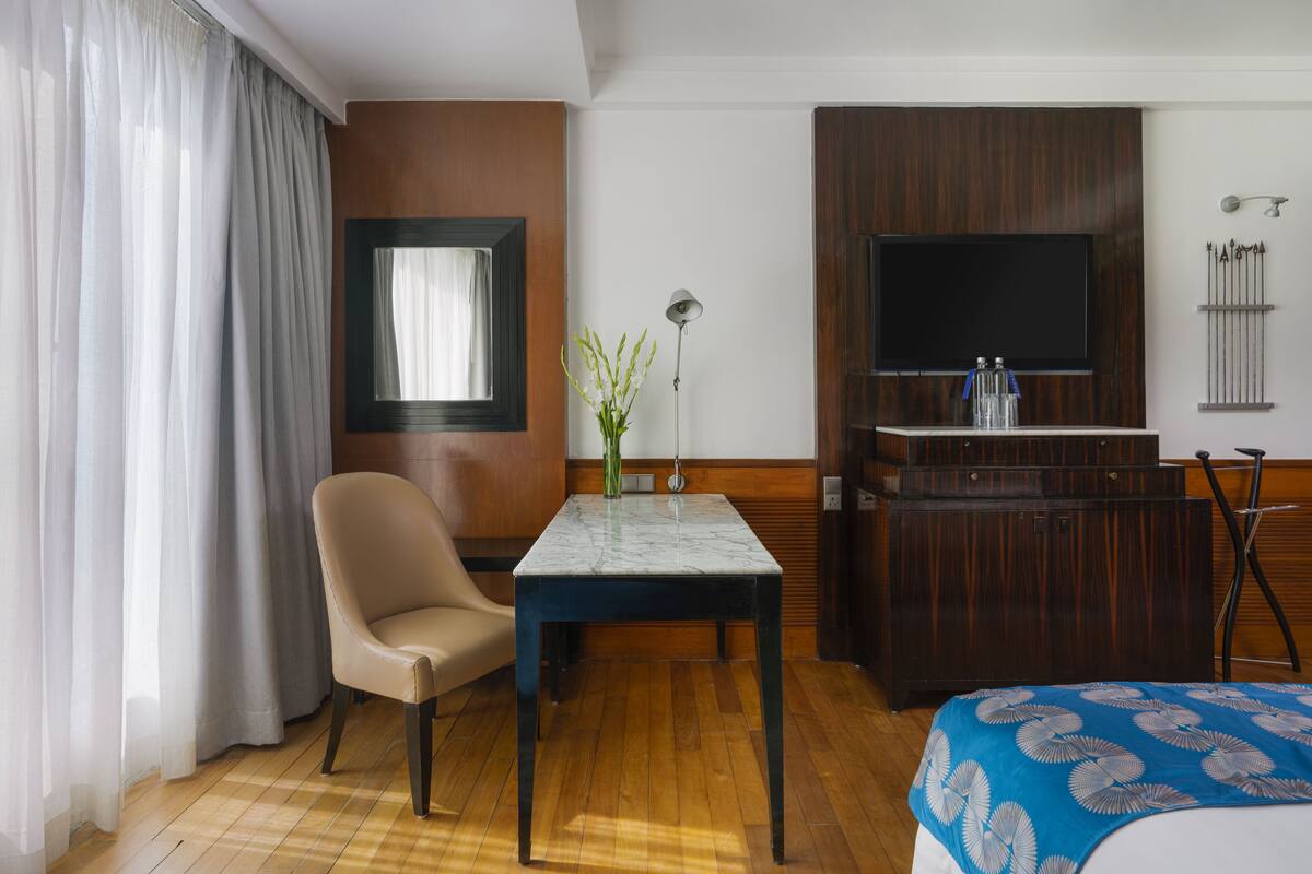 A hotel room with a wooden floor, a bed with a blue floral bedspread, a table with a vase, a chair, a TV on a wooden cabinet, and a window with curtains.