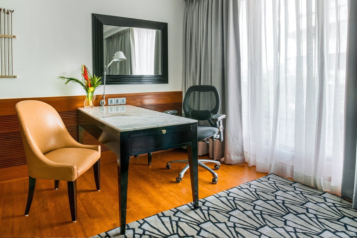 A hotel room with a wooden floor, a desk with a chair, a mirror, a lamp, and a window with curtains.