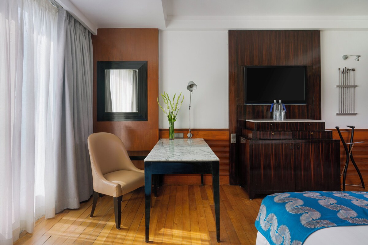 A hotel room with a wooden floor, a bed with a blue floral bedspread, a table with a vase, a chair, a TV on a wooden cabinet, and a window with curtains.