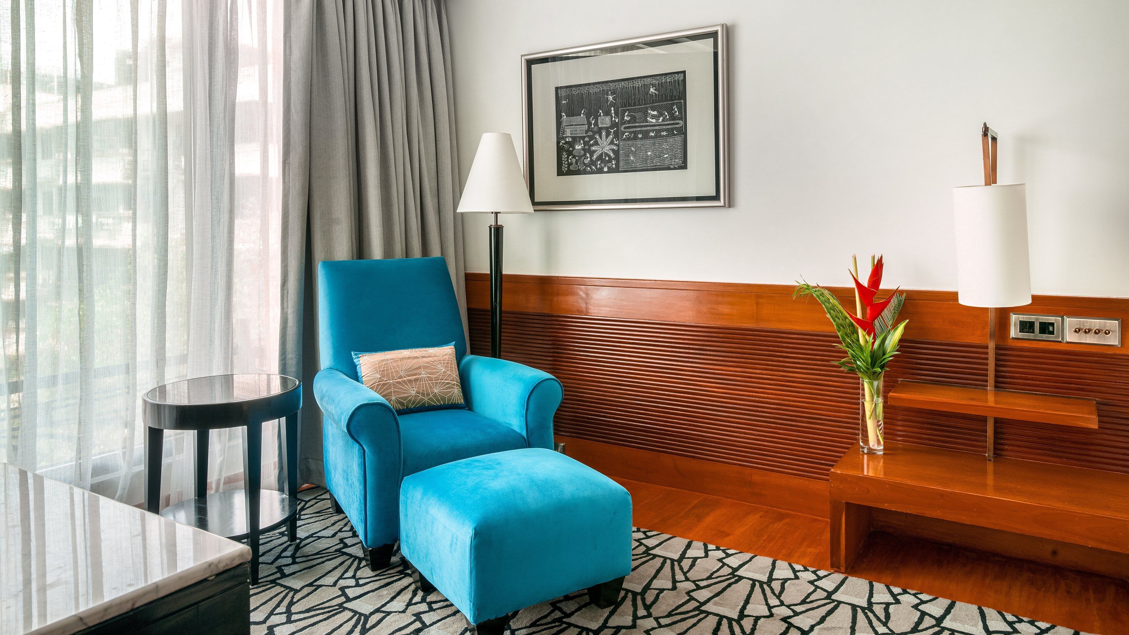 A hotel room with a blue armchair, a small table, a bench, a framed picture on the wall, and a vase with flowers.