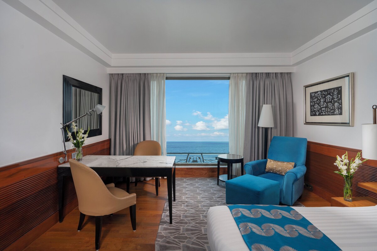 A hotel room with a bed, a desk, a chair, a sofa, a small table, and a view of the sea.