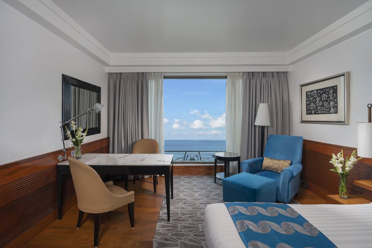 A hotel room with a bed, a desk, a chair, a sofa, a small table, and a view of the sea.