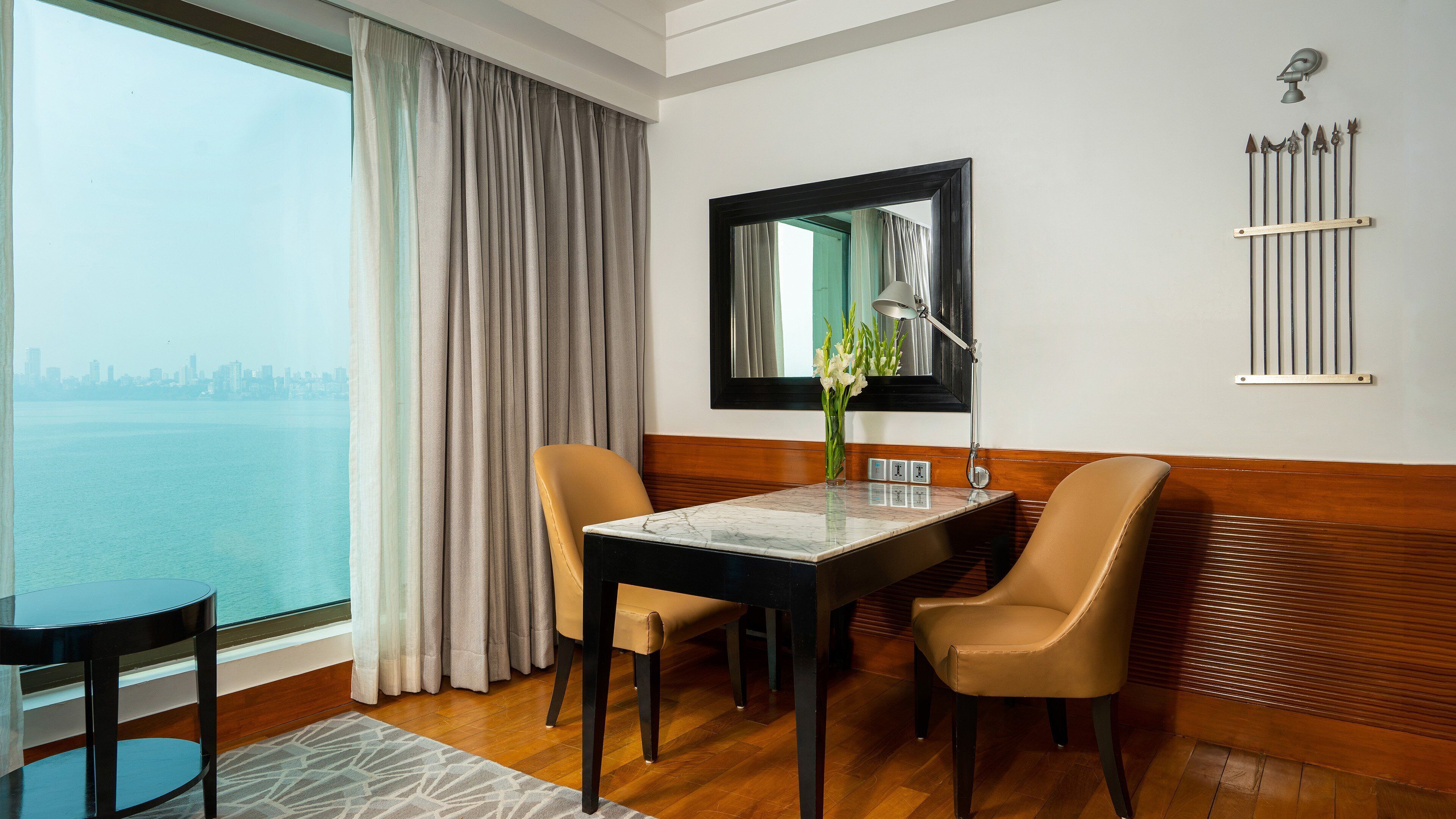 A hotel room with a dining table, two chairs, a mirror, a lamp, and a view of the city skyline.
