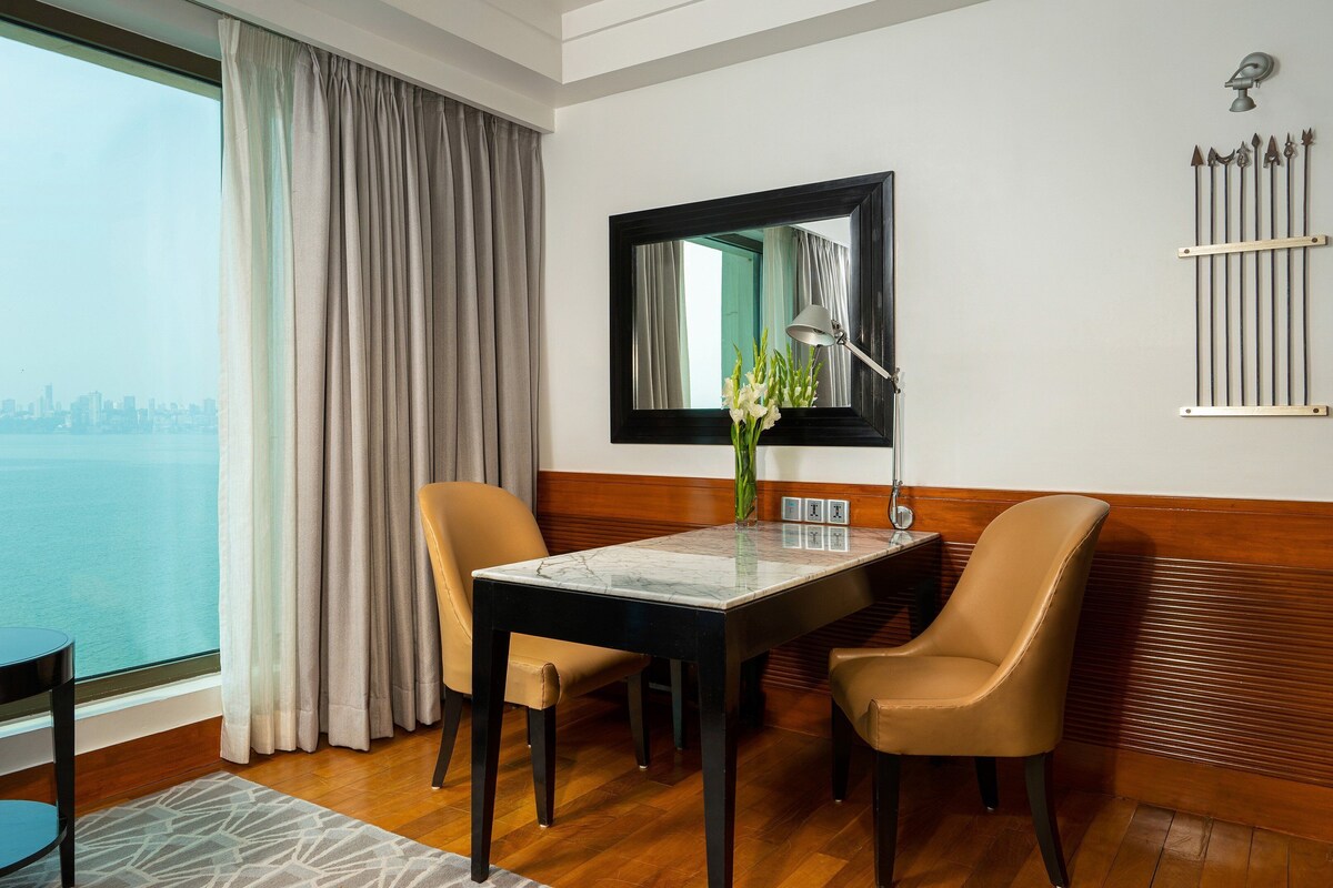 A hotel room with a dining table, two chairs, a vase with flowers, a mirror, and a view of the city skyline.