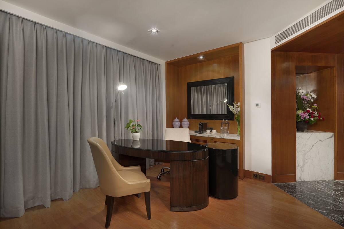A modern hotel room with a wooden dining table, a beige chair, a small potted plant, a coffee machine, and a marble countertop.