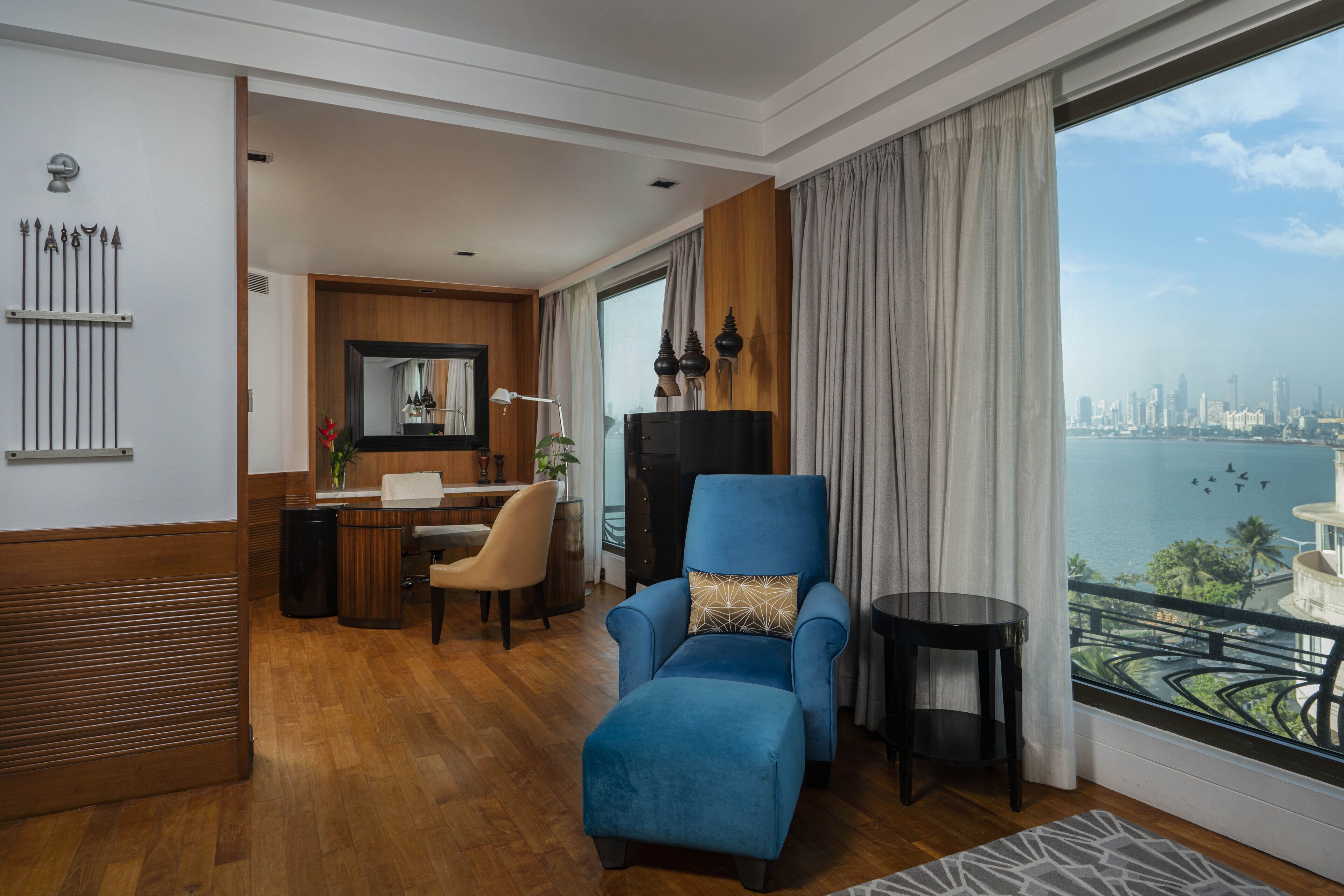 A modern hotel room with a large window offering a city view, a blue armchair, a dining area, and a wooden accent wall.