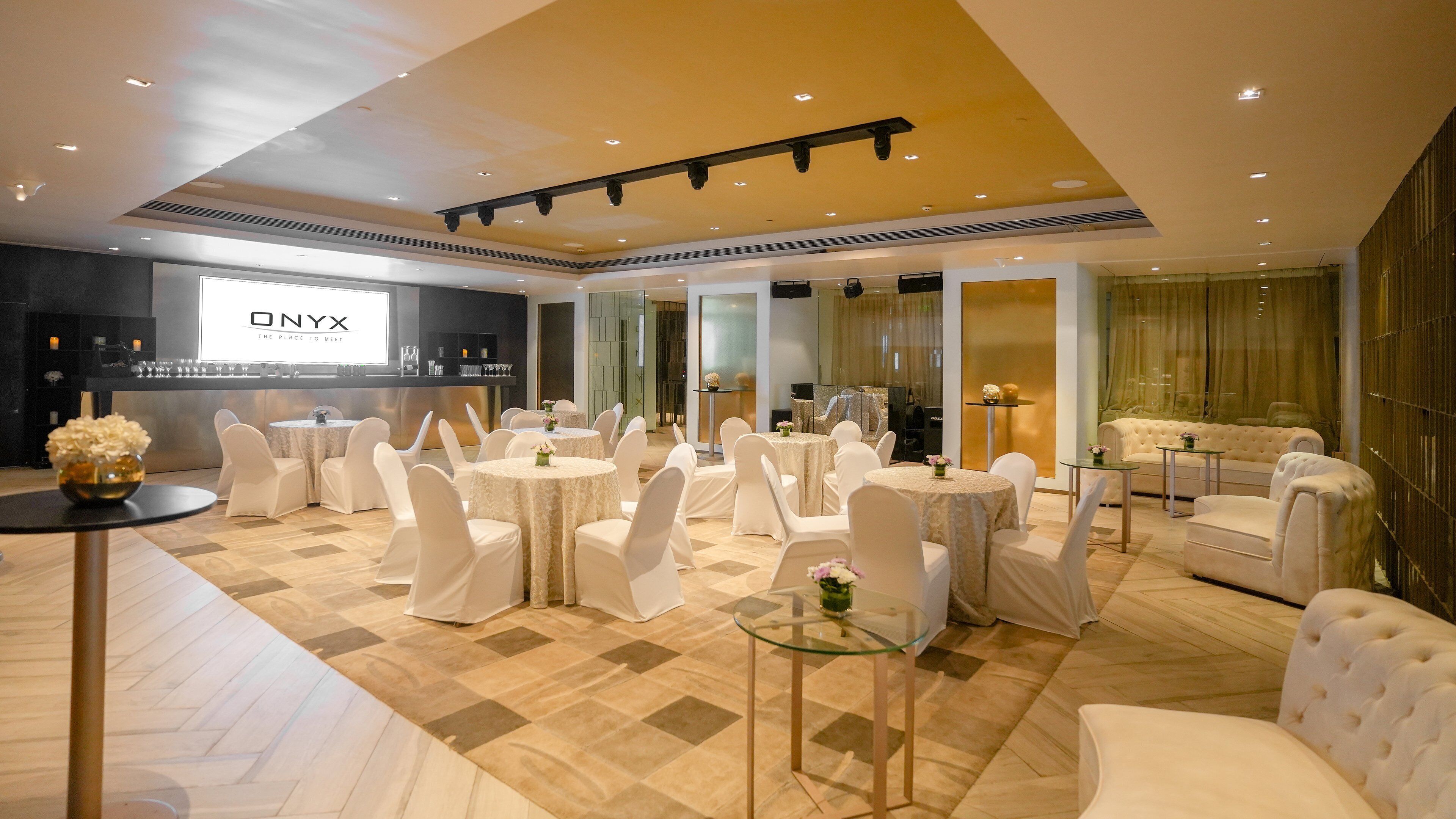 Une salle de réception spacieuse, dotée de tables rondes et de chaises blanches, un espace d’accueil avec un grand écran et un canapé confortable.