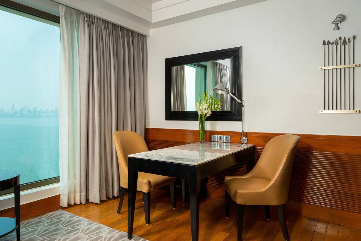 A hotel room with a dining table, two chairs, a vase with flowers, a mirror, and a view of the city skyline.