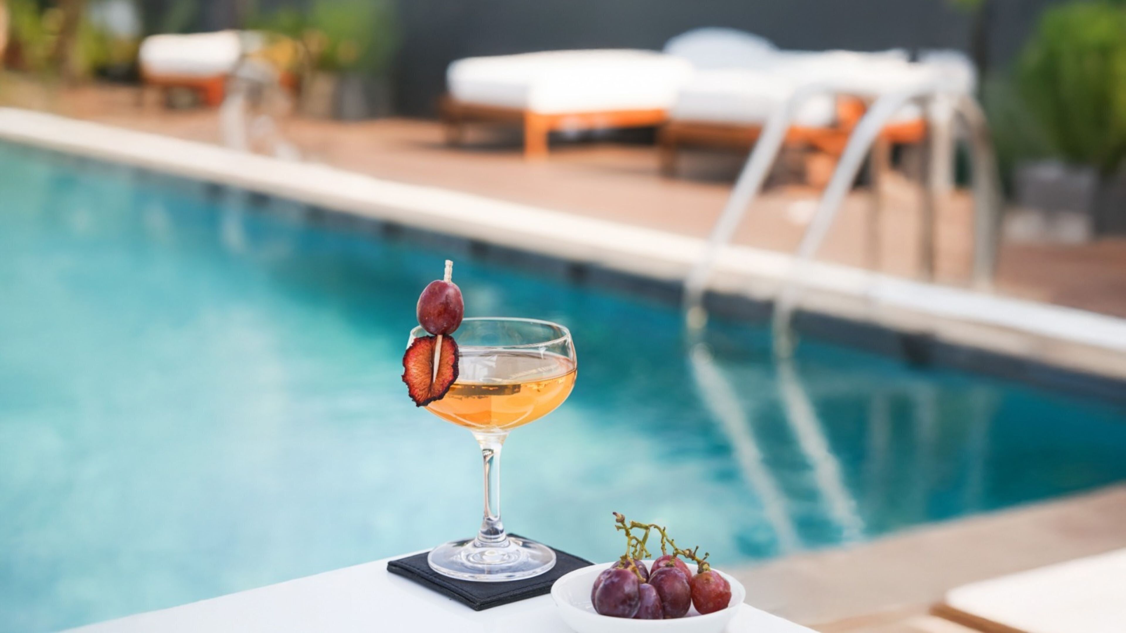 Escena junto a la piscina con un vaso de bebida, un plato de uvas y sillas de descanso.