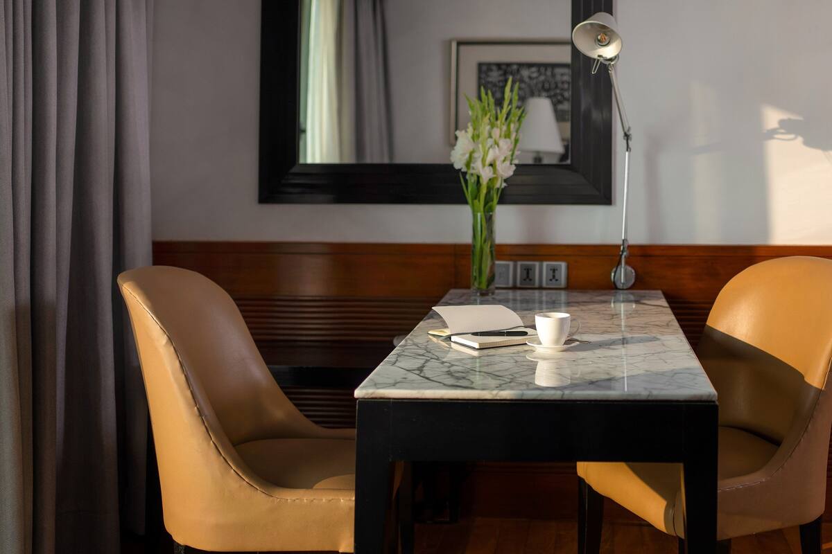 A hotel room with a wooden panel wall, a marble table, two leather chairs, a white coffee cup, a notebook, a pen, a lamp, and a vase with flowers.