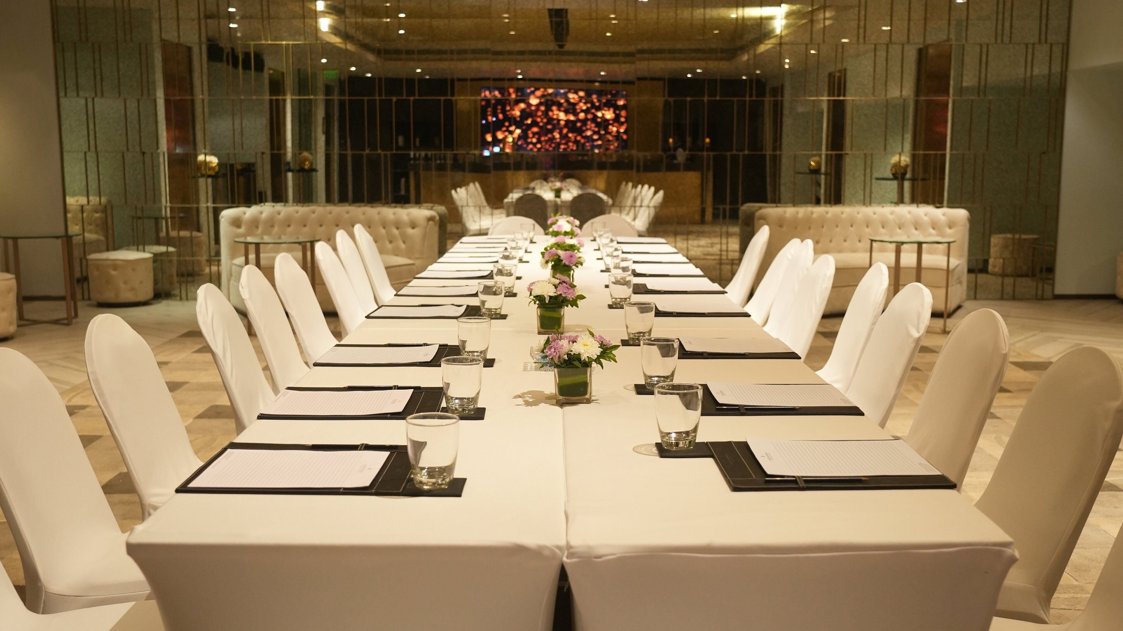 A long conference table with white chairs, glassware, and floral centerpieces, set in a modern room with a large window and a decorative wall panel.