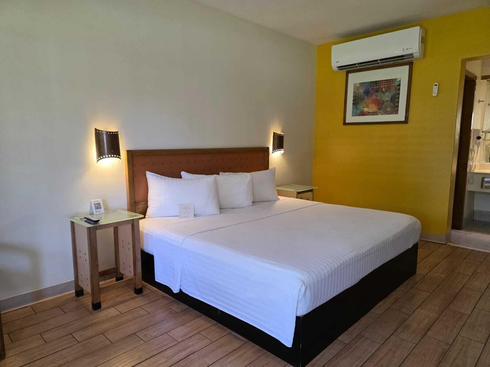Una habitación de hotel con una cama grande, dos mesitas de noche, un equipo de aire acondicionado y un cuadro en la pared.