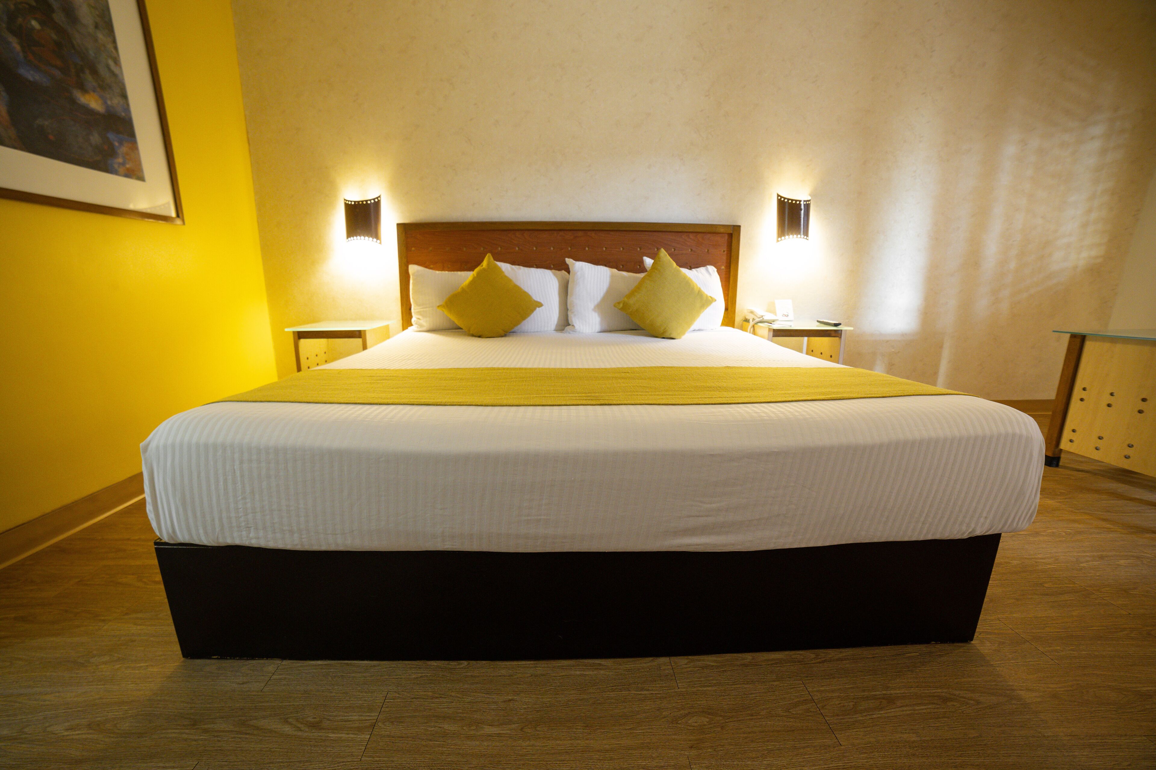 Uma cama grande com cabeceira de madeira, duas mesas de cabeceira e uma parede em tom de amarelo.