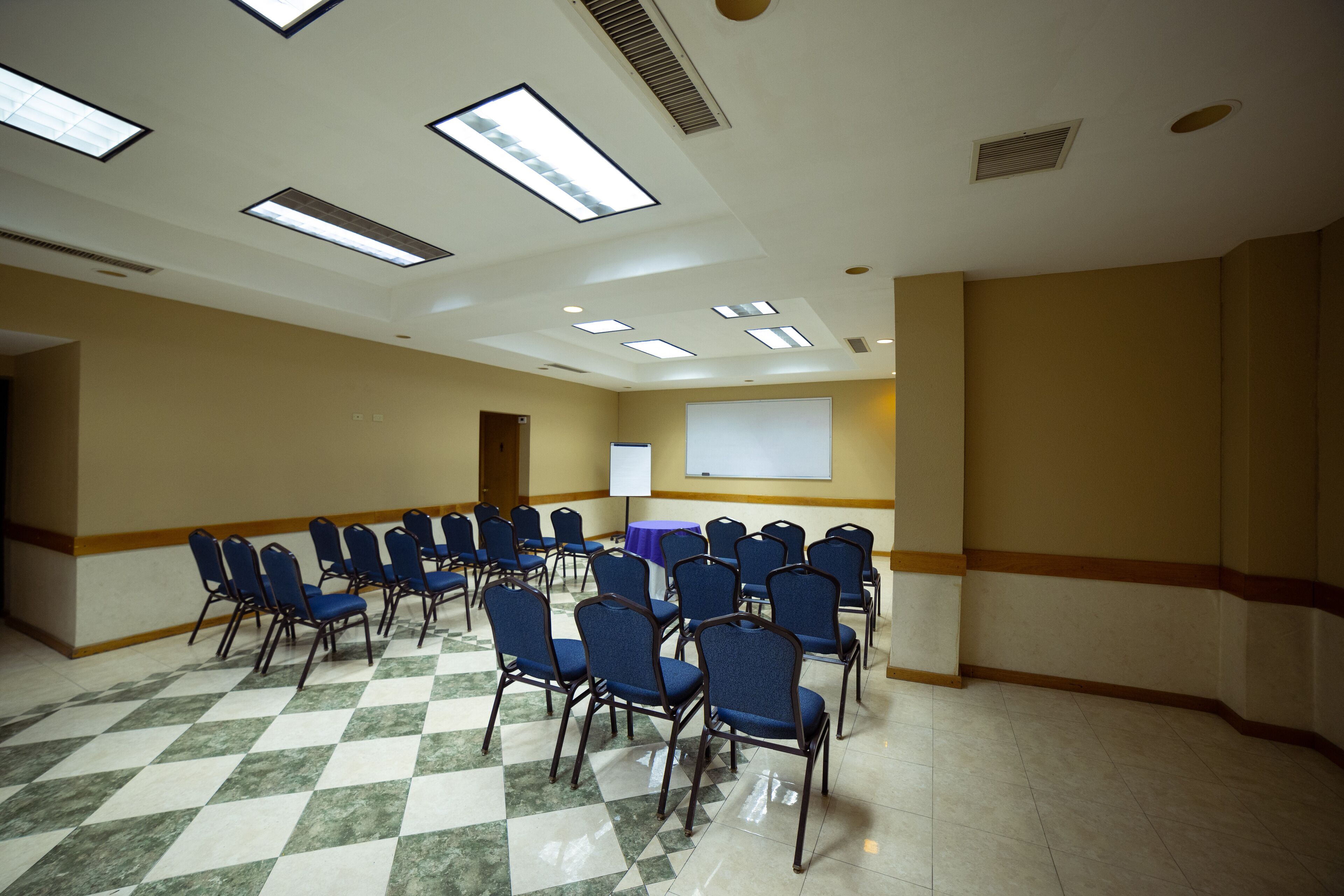 Un salón de conferencias con suelo de cuadros, filas de sillas azules, una pantalla de proyector y una mesa con mantel morado.