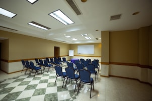 Un salón de conferencias con suelo de cuadros, filas de sillas azules, una pantalla de proyector y una mesa con mantel morado.