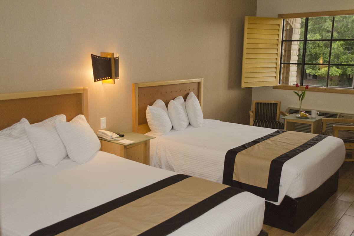 Habitación de hotel con dos camas, una mesita de noche con lámpara, una silla, una pequeña mesa con un jarrón y una taza, y una ventana con persianas.
